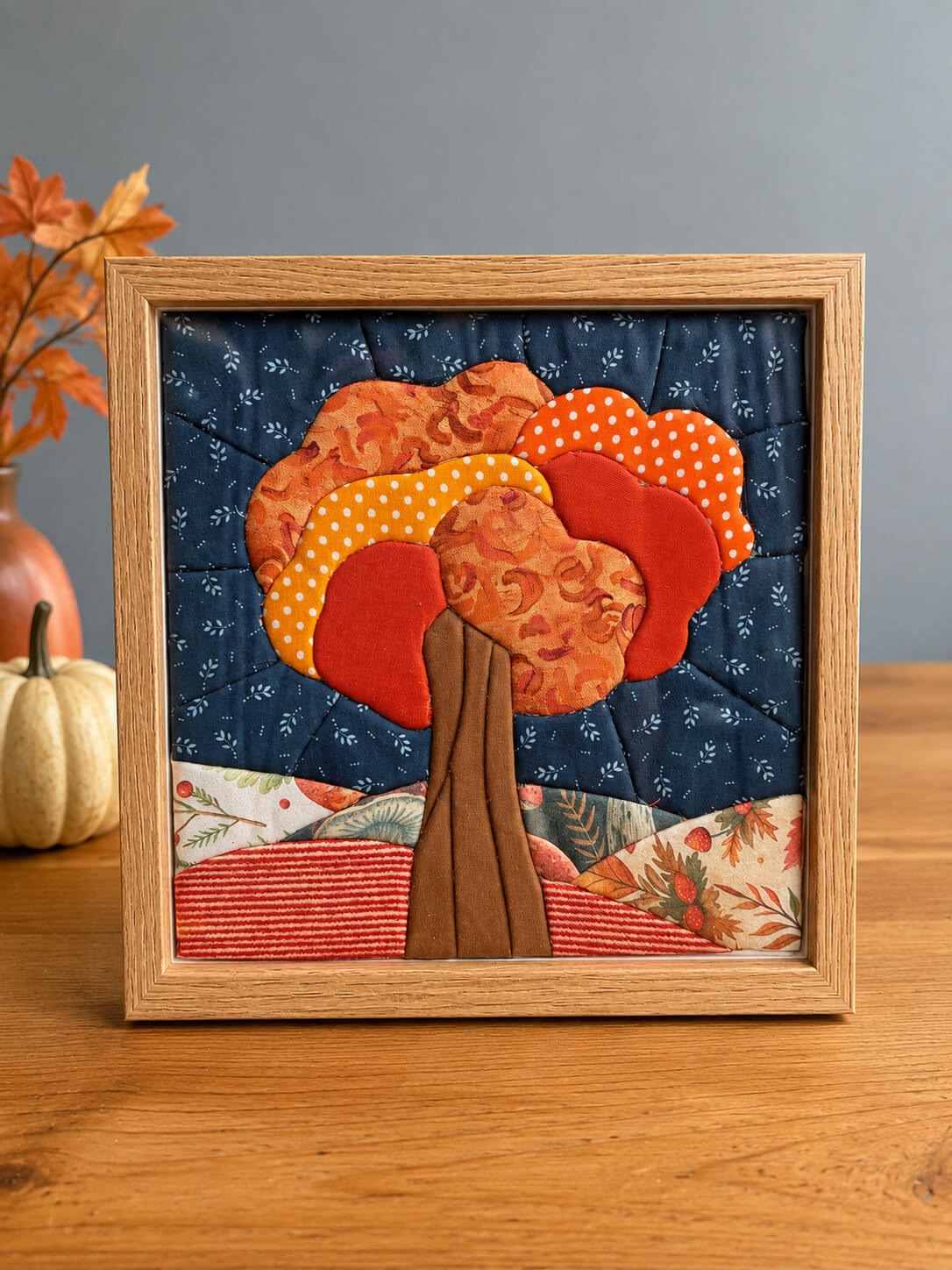 Kinusaiga tree in a natural wood frame, standing on a table with a brown trunk and a canopy of orange, red and yellow fabric, against a navy sky, over rolling hills of red stripe and autumn botanical prints.
