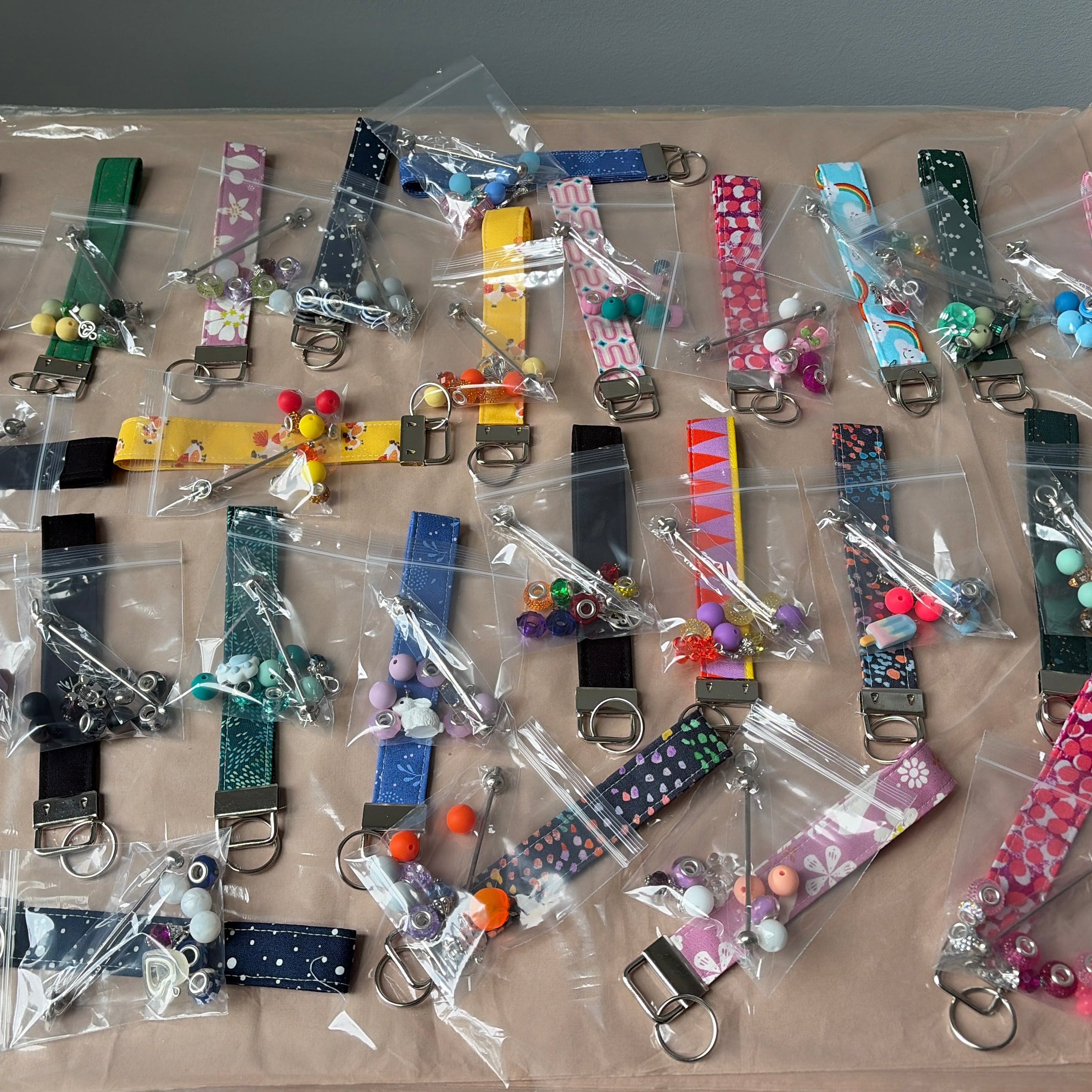 A collection of colorful fabric keychains and small accessories, each individually sealed in clear plastic bags, arranged on a tan display board.
