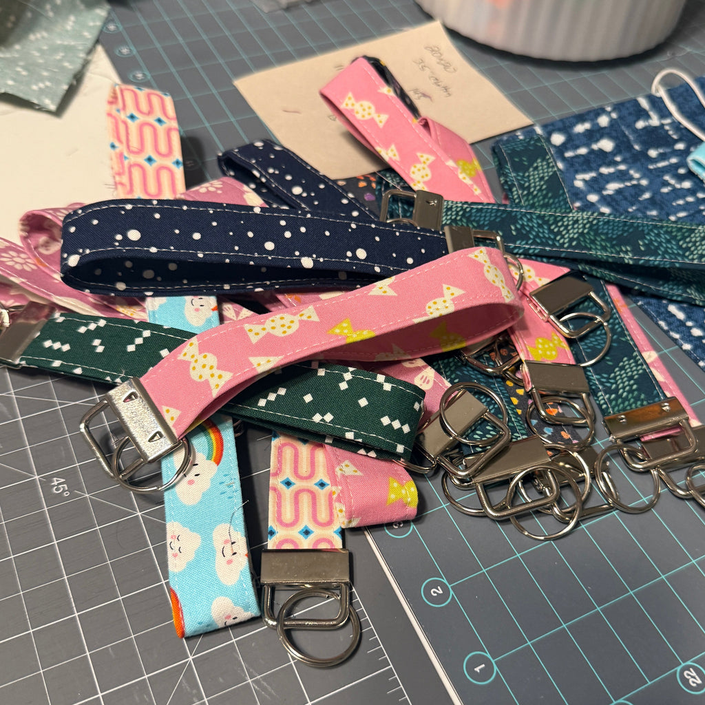 A pile of colorful fabric keychains with metal keyrings and buckles spread out on a gray cutting mat at a craft table. The keychains feature various patterns including stars, cactus prints, and abstract designs in pink, blue, green, and navy.