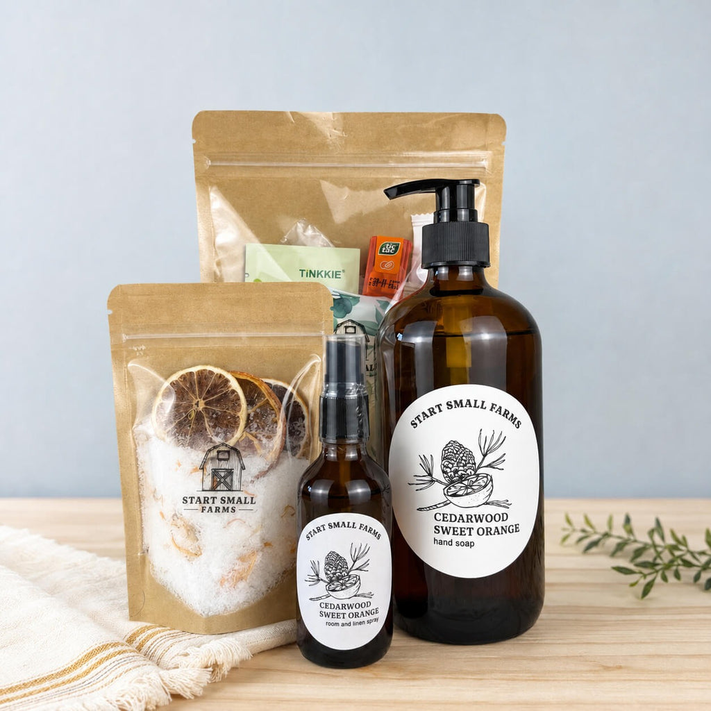 The After the Bell gift set on a wood surface: a kraft pouch of citrus bath soak with dried orange slices and calendula, an amber Cedarwood Sweet Orange hand soap pump, a matching room and linen spray, and the Break Glass essentials pouch behind