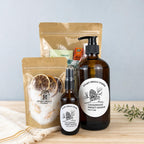 The After the Bell gift set on a wood surface: a kraft pouch of citrus bath soak with dried orange slices and calendula, an amber Cedarwood Sweet Orange hand soap pump, a matching room and linen spray, and the Break Glass essentials pouch behind