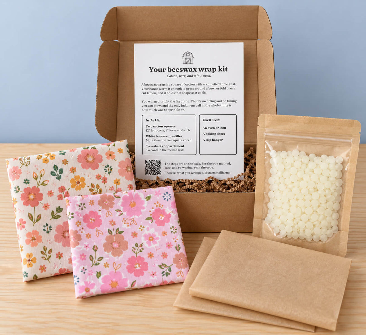 A beeswax wrap kit displayed in an open kraft cardboard box on a wooden surface against a light blue background. The kit includes two pieces of fabric, a clear bag filled with white wax melts, parchment paper, and an instruction card inside the box.