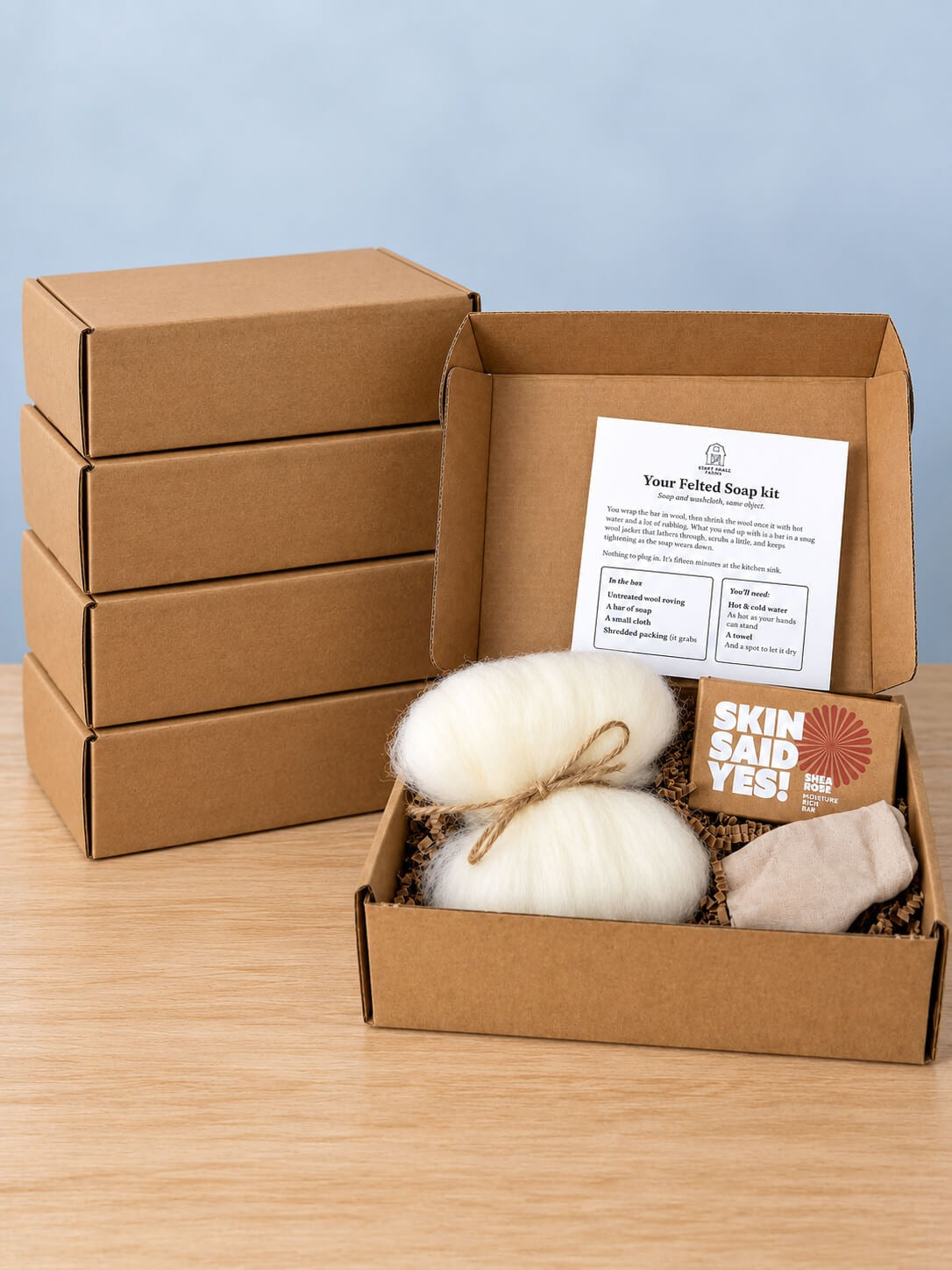 Five Felted Wool Soap DIY Kit boxes, four stacked closed and one open showing a bundle of cream wool roving tied with twine, a boxed bar of soap, a folded cloth and the kit card standing in the lid
