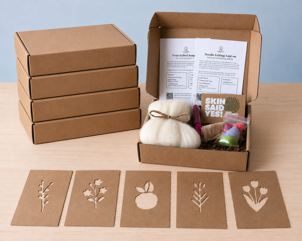 Five kit boxes, four stacked and one open with both instruction cards, a wool-wrapped bar, a felting needle and a bag of colored roving, with five kraft stencil cards laid out in front: a sprig, a flower cluster, an apple, a fern and tulips
