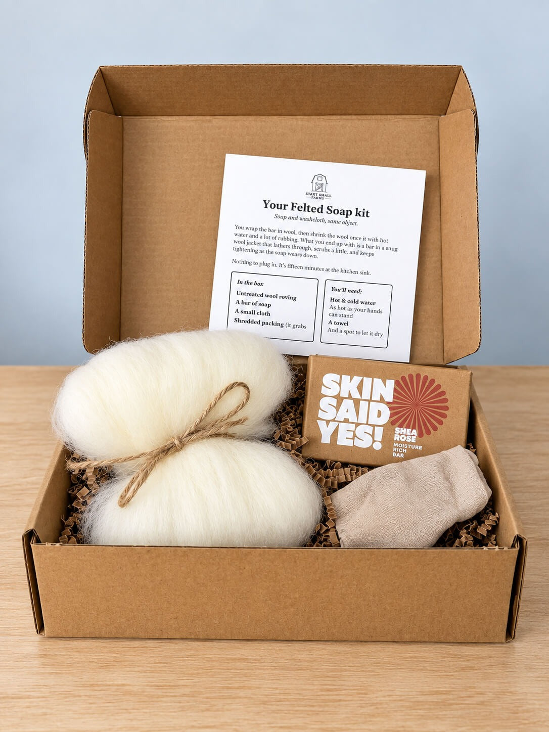 An open kraft box with crinkle paper holding a bundle of cream wool roving tied with twine, a boxed bar of soap and a folded cloth, with the Start Small Farms kit card standing in the lid