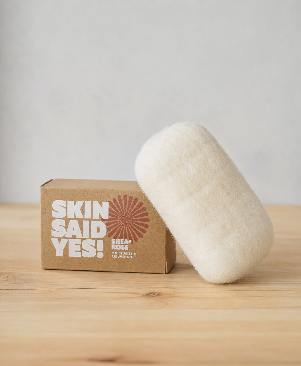 A finished bar in its cream wool jacket leaning against the soap's kraft box on a pine table