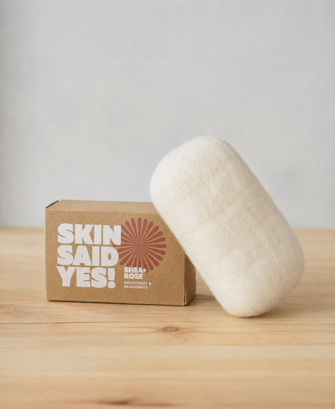 A finished bar in its cream wool jacket leaning against the soap's kraft box on a pine table