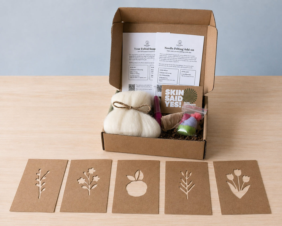 An open kraft box holding a wool-wrapped bar tied with twine, a felting needle, a bag of colored roving and both instruction cards, with five kraft stencil cards laid out in front of it: a sprig, a flower cluster, an apple, a fern and tulips