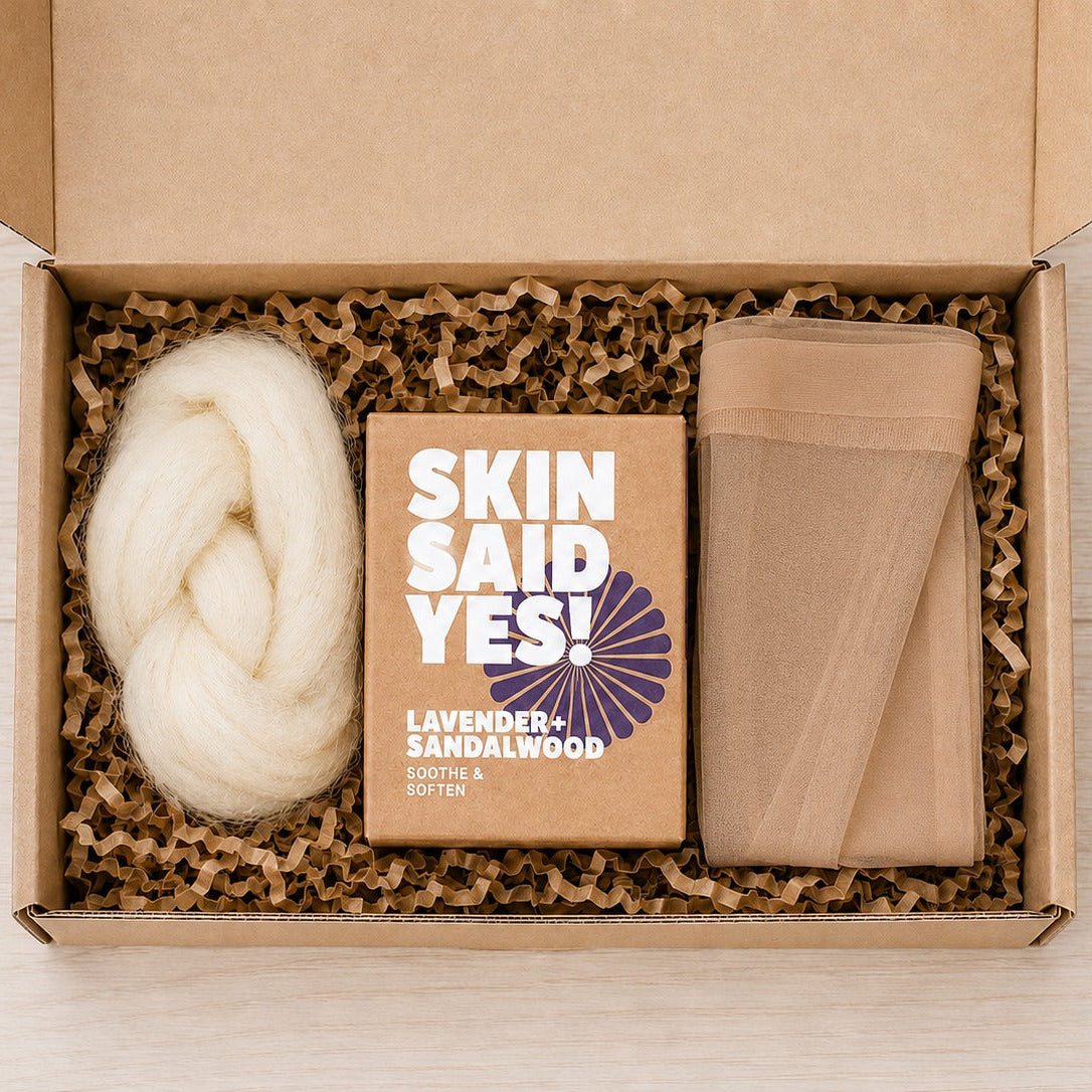 An open kraft box with crinkle paper holding a braid of cream wool roving, a boxed bar of soap, and a folded nylon stocking