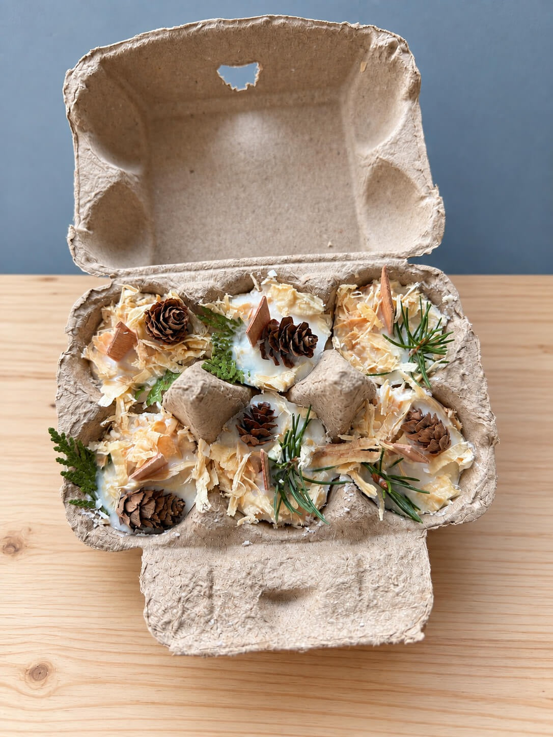 An open natural pulp egg carton on a wood table holding six round white wax firestarters, each topped with curls of pale wood shavings, a small pinecone, a chip of wood and a sprig of evergreen.