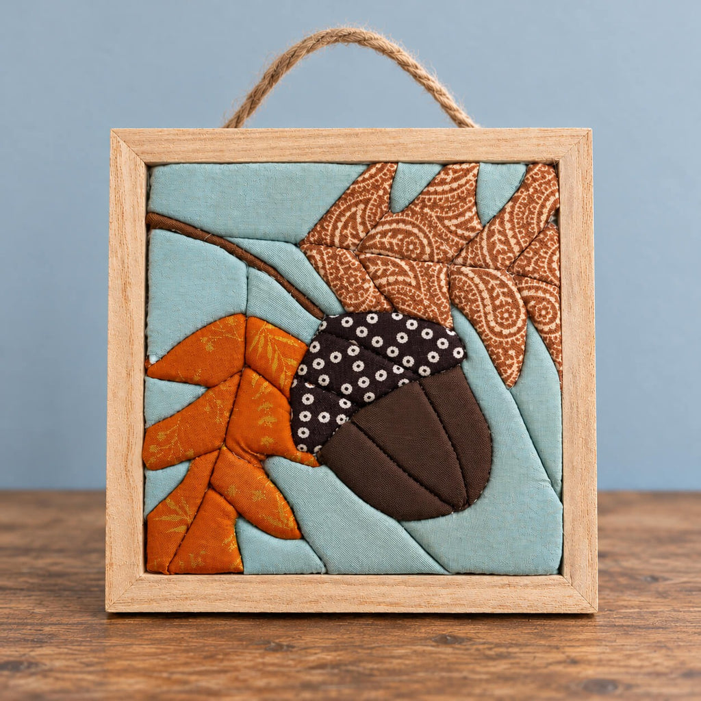 A small square kinusaiga wall hanging of an acorn in a natural wood frame with a rope hanger, standing on a wood table against a blue wall: a brown acorn with a dark polka dot cap, a burnt orange oak leaf and a brown paisley oak leaf on a branch, against a soft blue background.