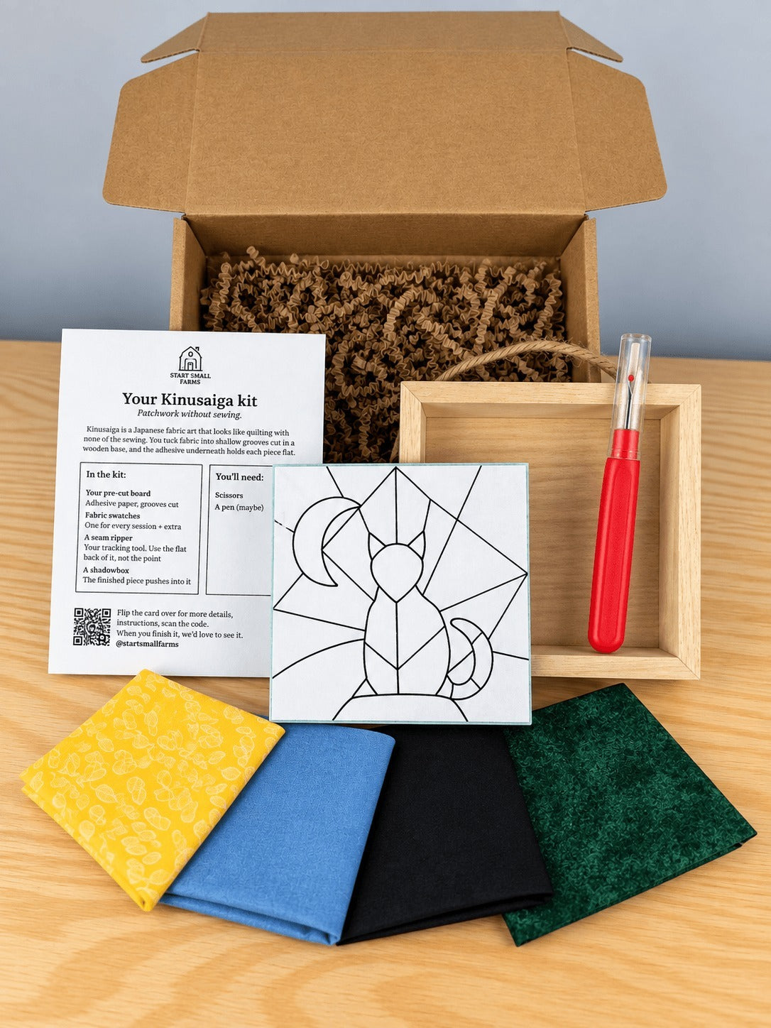 Everything in the kit, unboxed on a wood table: the open kraft shipping box with crinkle paper, the kit card, the pre-grooved board printed with the cat and crescent moon outline, an empty natural wood shadowbox with a twine hanger, a seam ripper, and four fabric swatches in yellow print, blue, black and green.