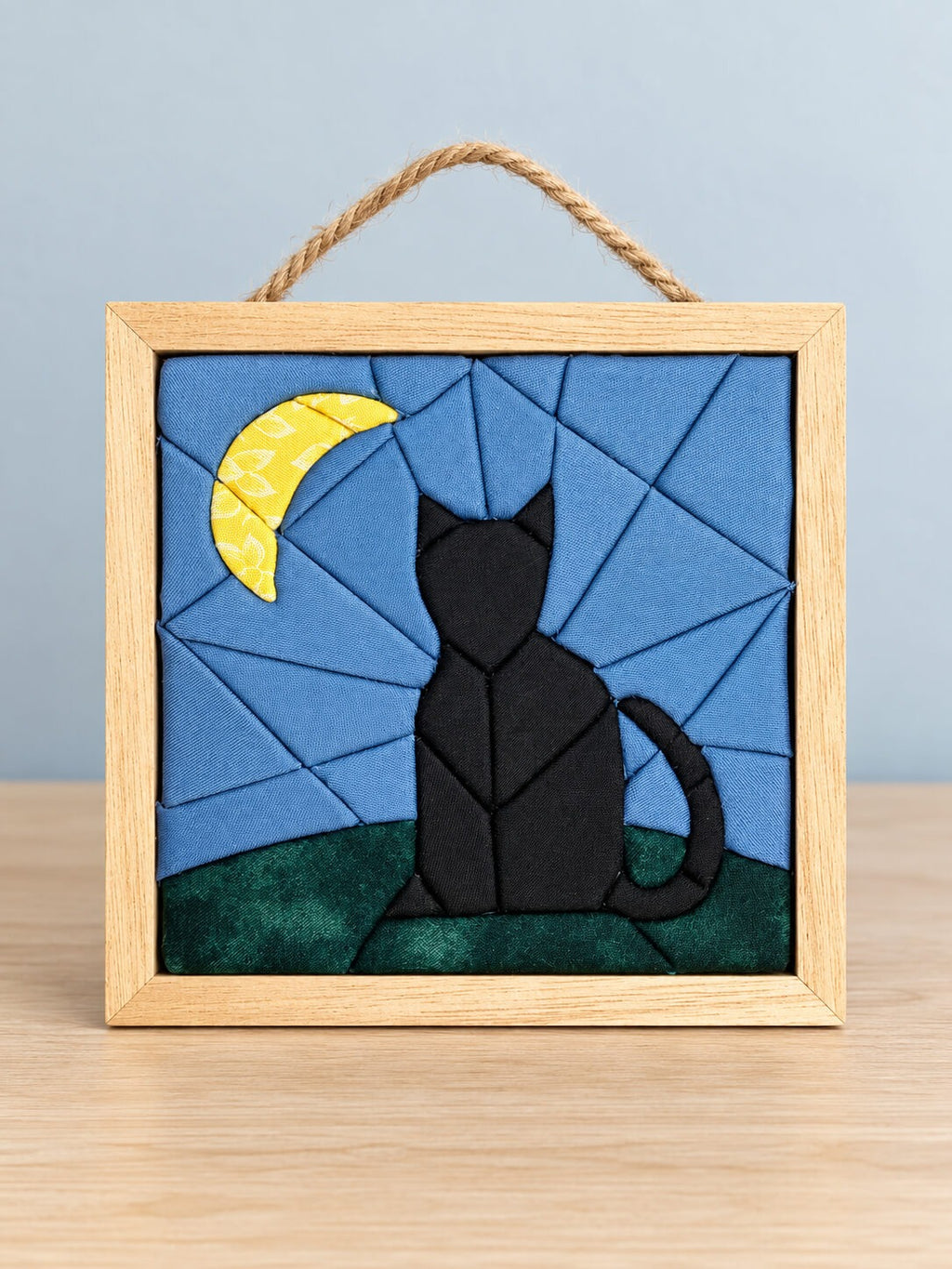 A small square kinusaiga wall hanging in a natural wood frame with a twine hanger, standing on a wood table: a black cat sitting with its tail curled, built from tucked fabric, on a green hill under a blue sky pieced in wedges, with a yellow floral crescent moon in the corner.