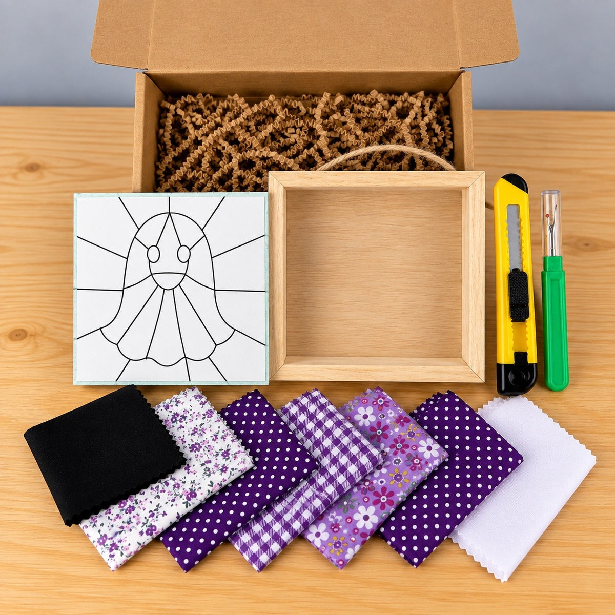 Everything in the kit on a wood table: the open kraft box with crinkle paper, the board printed with the ghost outline, an empty natural wood shadowbox with a twine hanger, a yellow craft knife, a green seam ripper, and seven fabric swatches in purples, lilac florals, gingham, black and white.