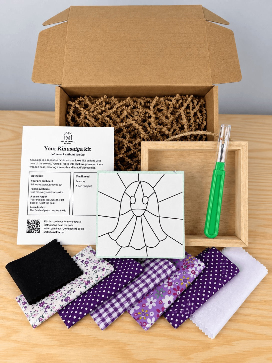Everything in the kit, unboxed on a wood table: the open kraft shipping box with crinkle paper, the kit card, the pre-grooved board printed with the ghost outline, an empty natural wood shadowbox with a twine hanger, a seam ripper, and seven fabric swatches in black, white and purple prints.