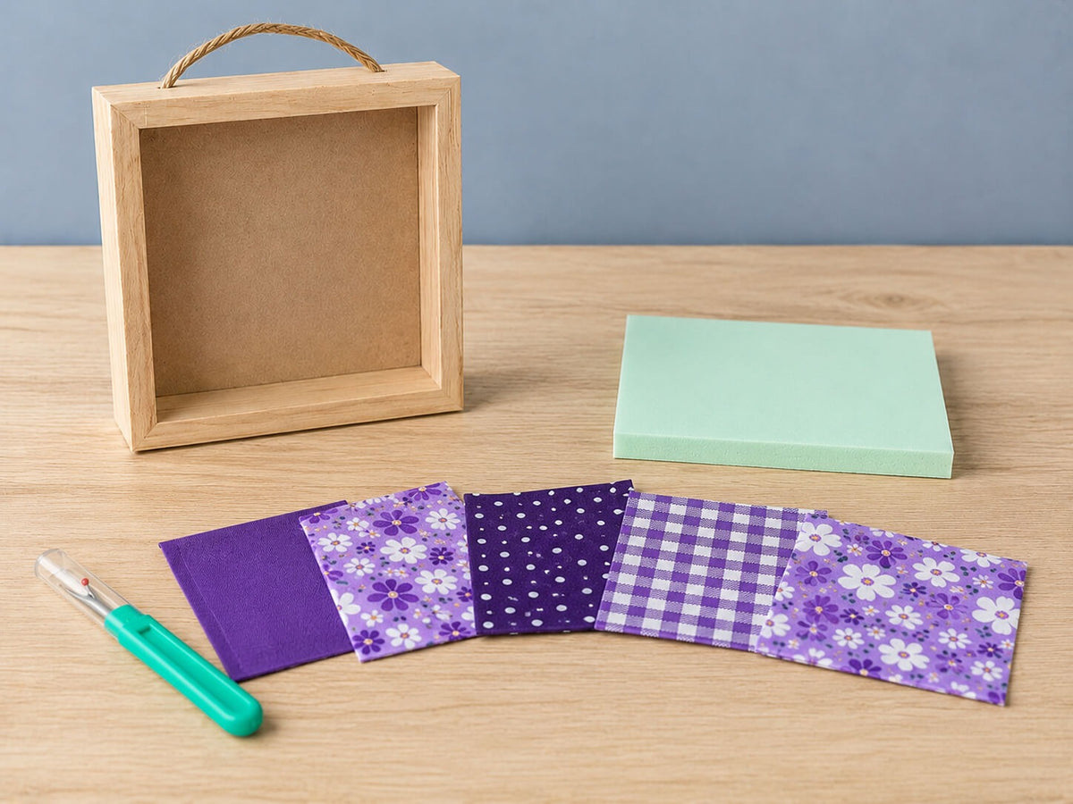 What comes in the kit, laid out on a wood table: an empty natural wood shadowbox with a twine hanger, a mint green pre-grooved foam board, a seam ripper, and a fan of six purple fabric swatches in solids, polka dots, gingham and florals.