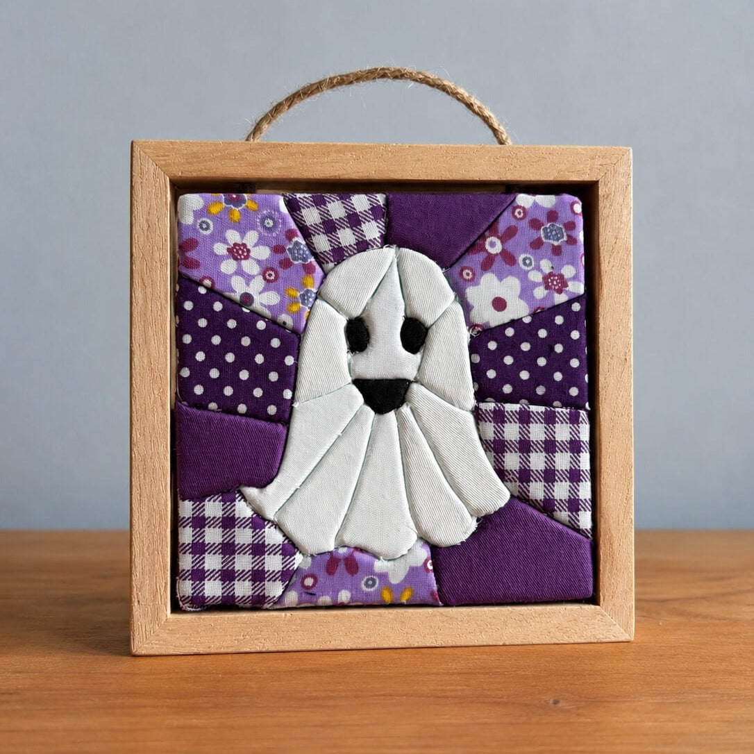 A small square kinusaiga wall hanging in a natural wood frame with a twine hanger: a white ghost with black eyes and an open mouth, built from tucked fabric, on a background of purple polka dot, gingham and floral patchwork.