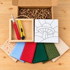 Everything in the kit on a wood table: the open kraft box with crinkle paper, an empty natural wood shadowbox with a twine hanger holding a red seam ripper and a yellow craft knife, the board printed with the mushroom outline, and six fabric swatches in peach print, light blue, red polka dot, two greens and cream.