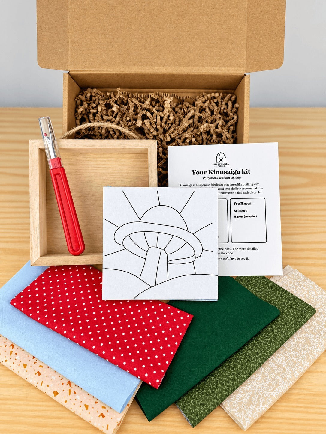 Everything in the kit, unboxed on a wood table: the open kraft shipping box with crinkle paper, the kit card, the pre-grooved board printed with the mushroom outline, an empty natural wood shadowbox with a twine hanger, a seam ripper, and six fabric swatches in red polka dot, light blue, greens and cream prints.