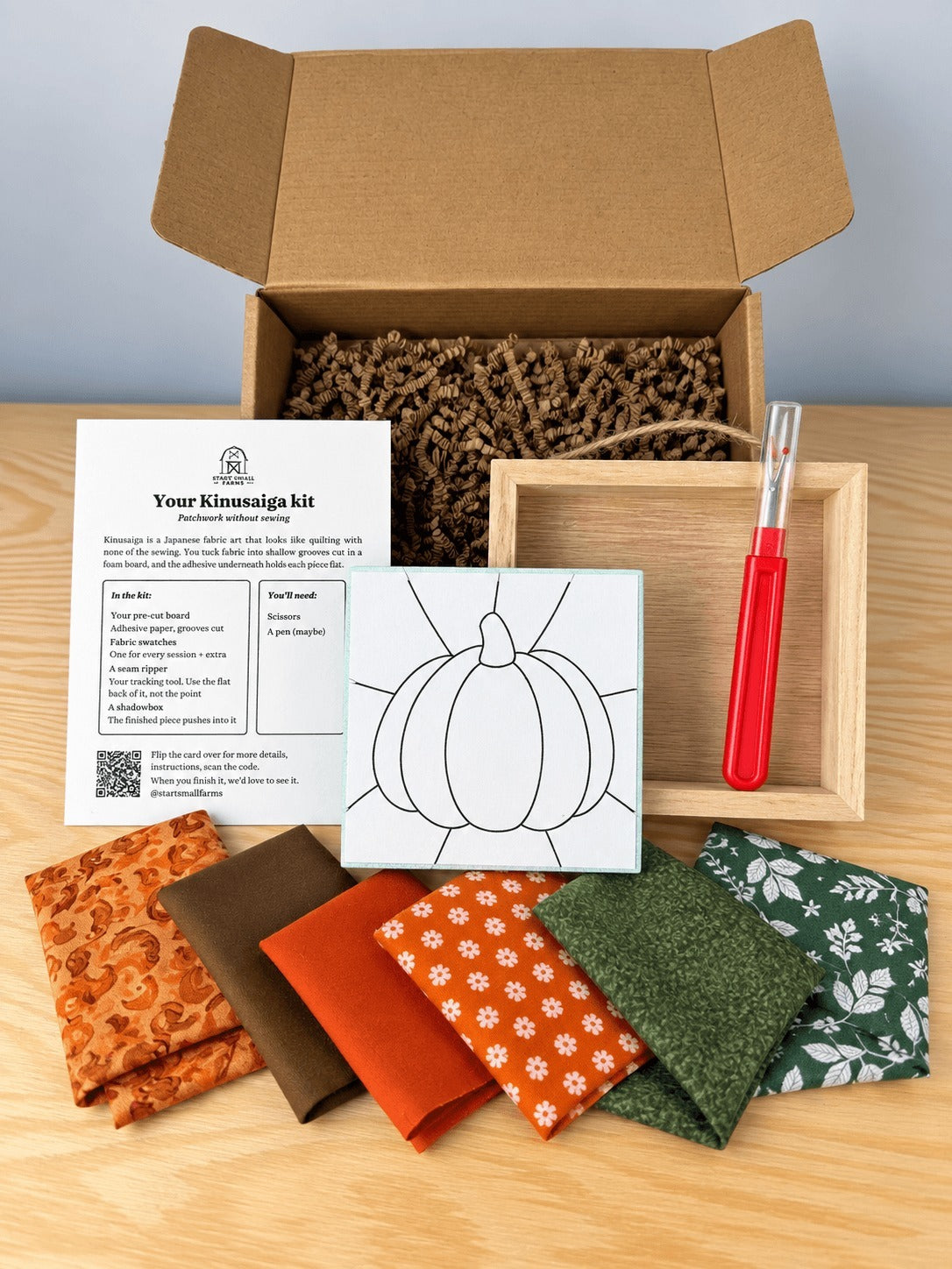 Everything in the kit, unboxed on a wood table: the open kraft shipping box with crinkle paper, the kit card, the pre-grooved board printed with the pumpkin outline, an empty natural wood shadowbox with a twine hanger, a seam ripper, and six fabric swatches in oranges, brown and greens.