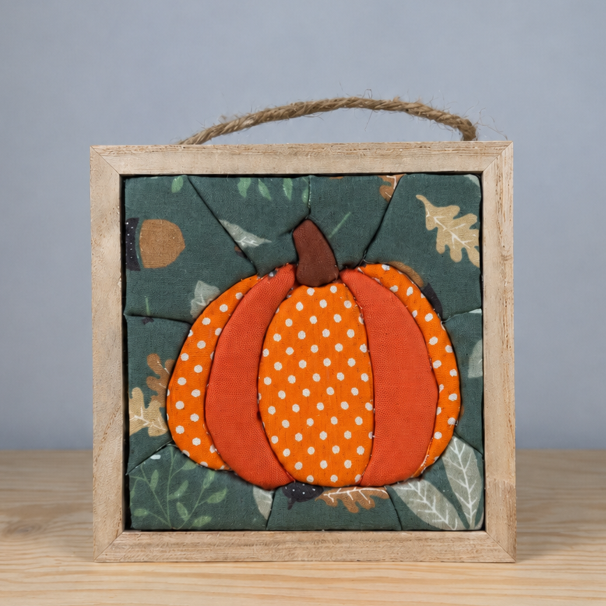 A square wooden hanging plaque featuring a stylized orange pumpkin with white polka dots on a dark teal autumn leaf background, displayed on a light wood surface against a gray backdrop.