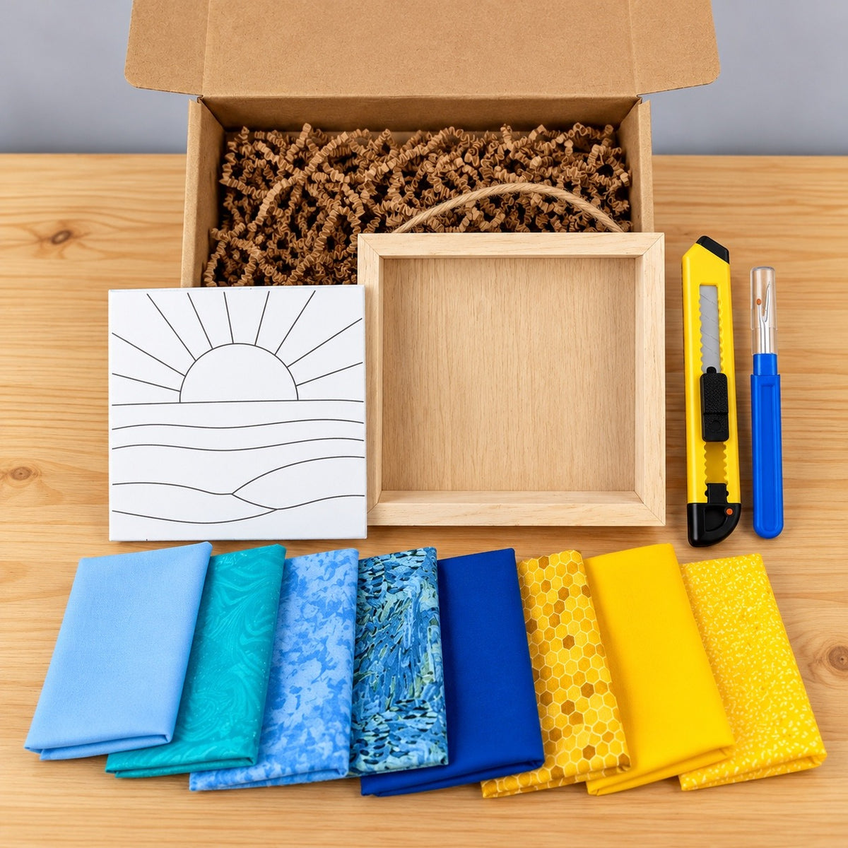 Everything in the kit on a wood table: the open kraft box with crinkle paper, the board printed with the sun and waves outline, an empty natural wood shadowbox with a twine hanger, a yellow craft knife, a blue seam ripper, and eight fabric swatches in blues, teals and yellows, one with a honeycomb print.