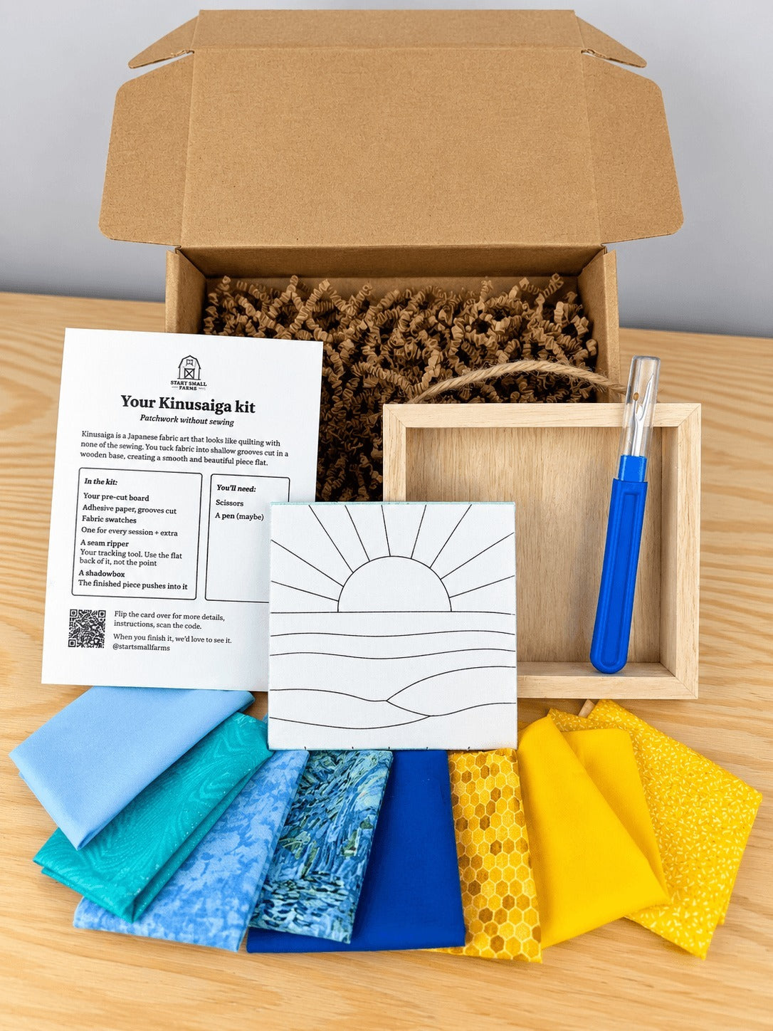 Everything in the kit, unboxed on a wood table: the open kraft shipping box with crinkle paper, the kit card, the pre-grooved board printed with the sun and waves outline, an empty natural wood shadowbox with a twine hanger, a seam ripper, and eight fabric swatches in blues and yellows.