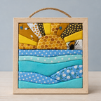 A square framed fabric art piece featuring a quilted sun and waves in yellow, blue, and teal fabrics, with a rope handle on top and a light wood frame, set against a neutral gray background.