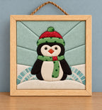 A small square kinusaiga wall hanging of a penguin in a natural wood frame with a rope hanger, against a blue-grey wall: a black and cream penguin with black glass eyes and an orange beak, wearing a red hat with a green polka dot brim and pompom and a matching green polka dot scarf, on a snowbank under a pale aqua sky pieced in wedges.