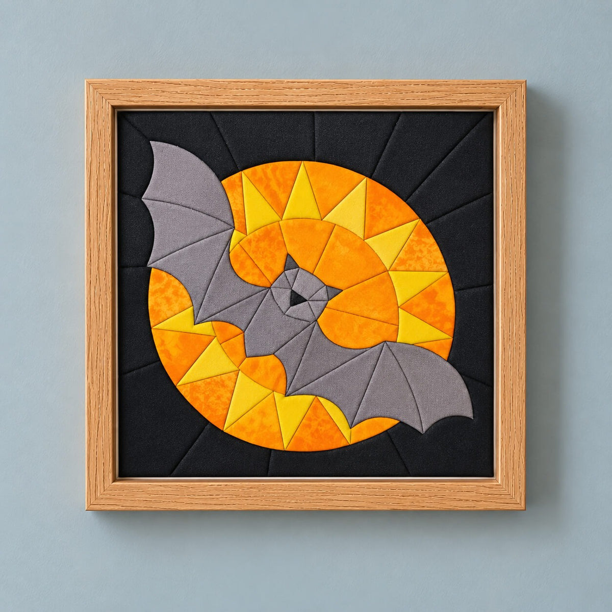 A square kinusaiga wall piece in a natural wood frame, hanging flat on a grey-blue wall: a grey bat with spread wings across a full moon of orange and yellow fabric triangles, on a black background of tucked fabric radiating out from the center.