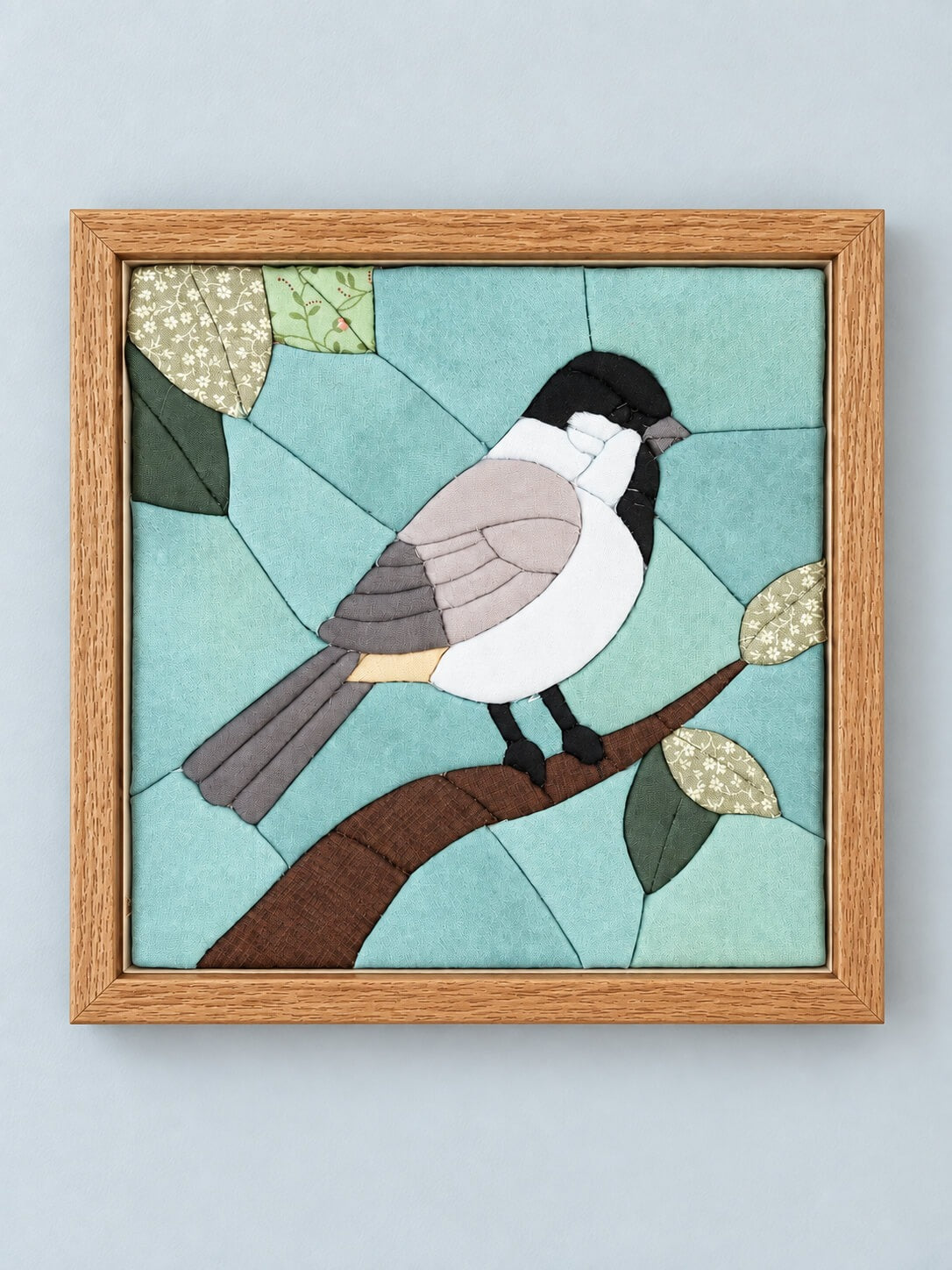 A square kinusaiga wall piece in a natural wood frame, hanging flat against a pale grey wall: a black-capped chickadee with a white cheek, grey wing and buff underside, perched on a brown branch with green floral-print leaves, on a sky of pieced soft blues and aquas.