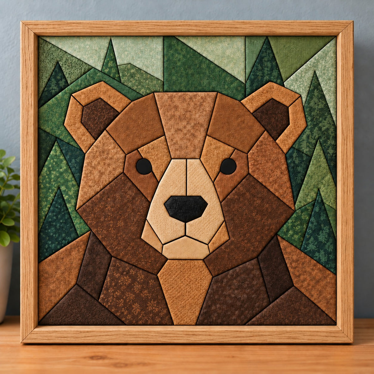 A chunky brown bear kinusaiga piece in a natural wood frame, standing on a wooden shelf against a blue-grey wall: a round geometric bear face in tan, caramel and chocolate brown fabrics, with a cream muzzle, black nose and dark inner ears, in front of pine trees pieced in five shades of green.