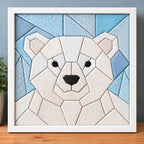 A chunky polar bear kinusaiga piece in a white frame, standing on a wooden shelf against a blue-grey wall: a round geometric bear face in white, ivory and cream fabrics, with pale pink inner ears, black eyes and a black nose, against a sky of icy blue triangles.