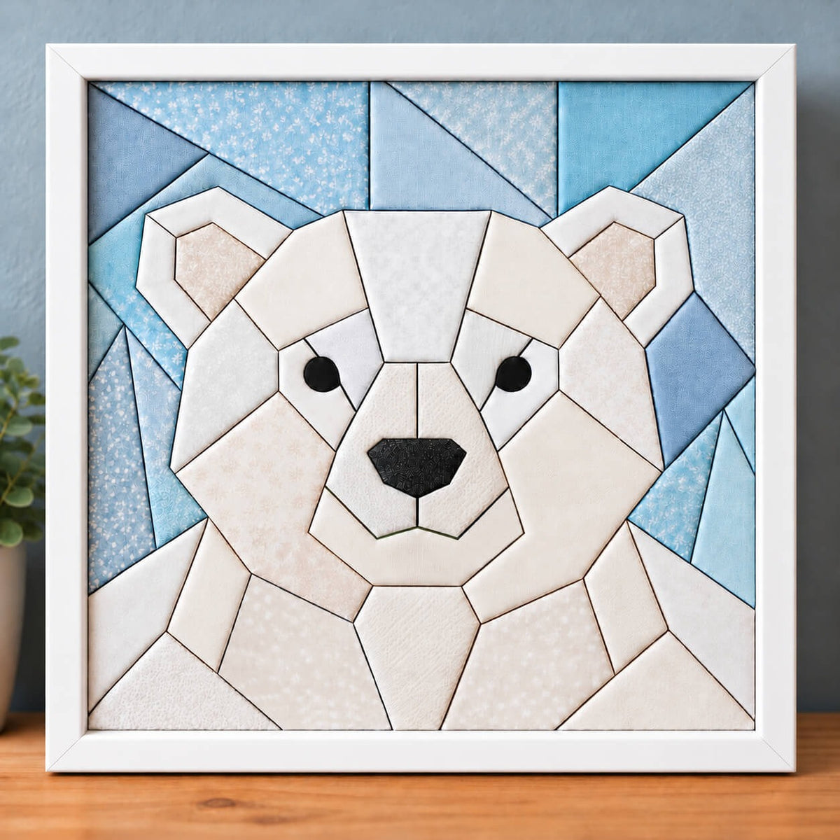 A chunky polar bear kinusaiga piece in a white frame, standing on a wooden shelf against a blue-grey wall: a round geometric bear face in white, ivory and cream fabrics, with pale pink inner ears, black eyes and a black nose, against a sky of icy blue triangles.