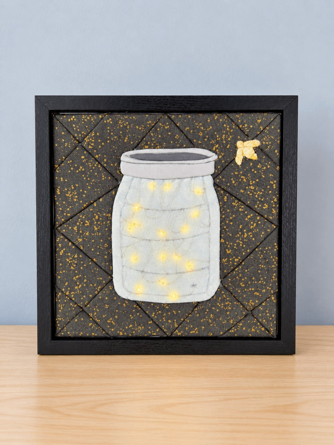 A square kinusaiga wall piece in a black frame, standing on a light wood table: a pale grey mason jar built from tucked fabric, lit from inside by a dozen small warm LED lights, on a background of dark grey fabric speckled with gold, pieced in triangles, with a small yellow fabric firefly in the top corner.
