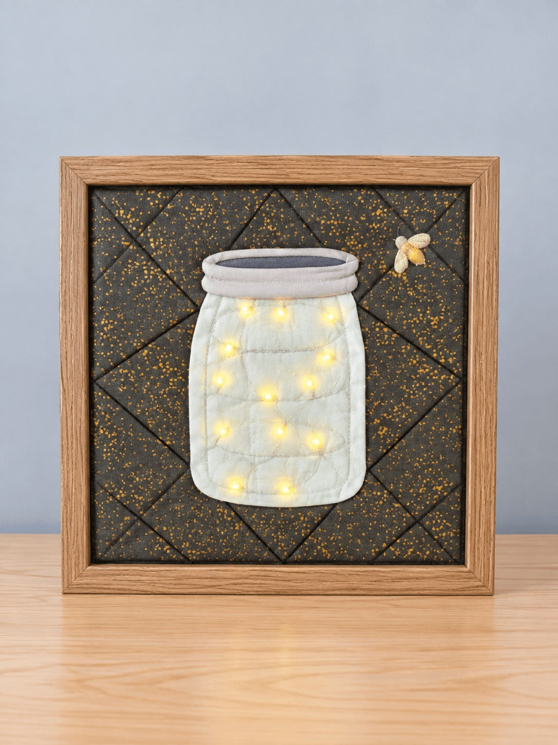 The same firefly jar design lit up, in a natural wood frame, standing on a light wood table.
