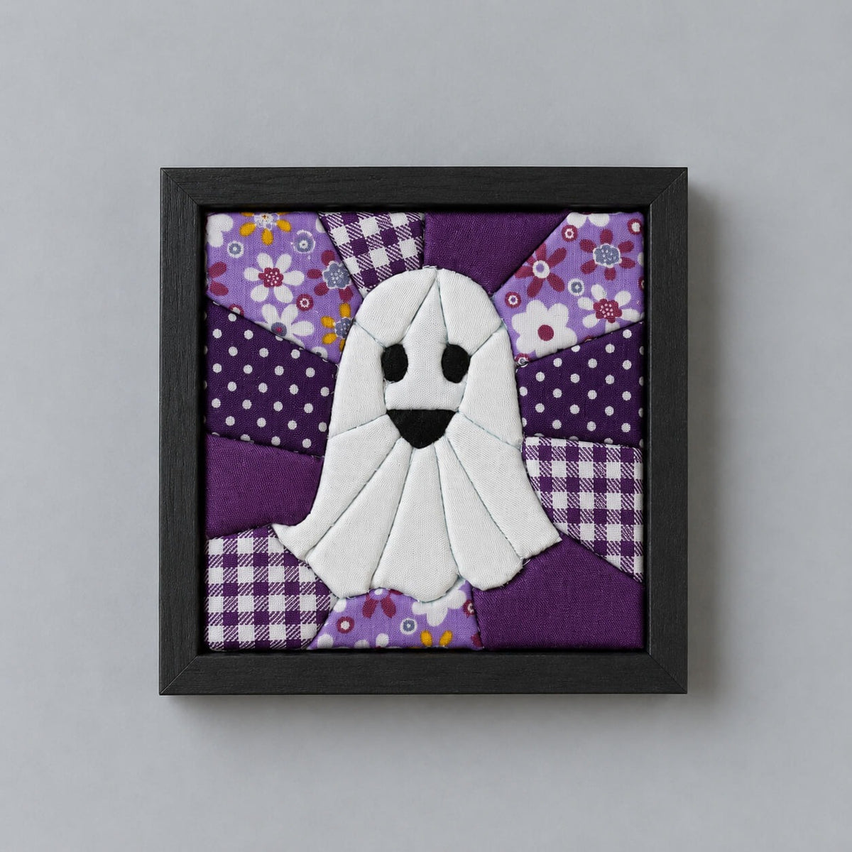 A square kinusaiga wall piece in a black wood frame: a white ghost with black eyes and an open mouth, built from tucked fabric, on a background of purple polka dot, gingham and floral patchwork radiating out from the center.