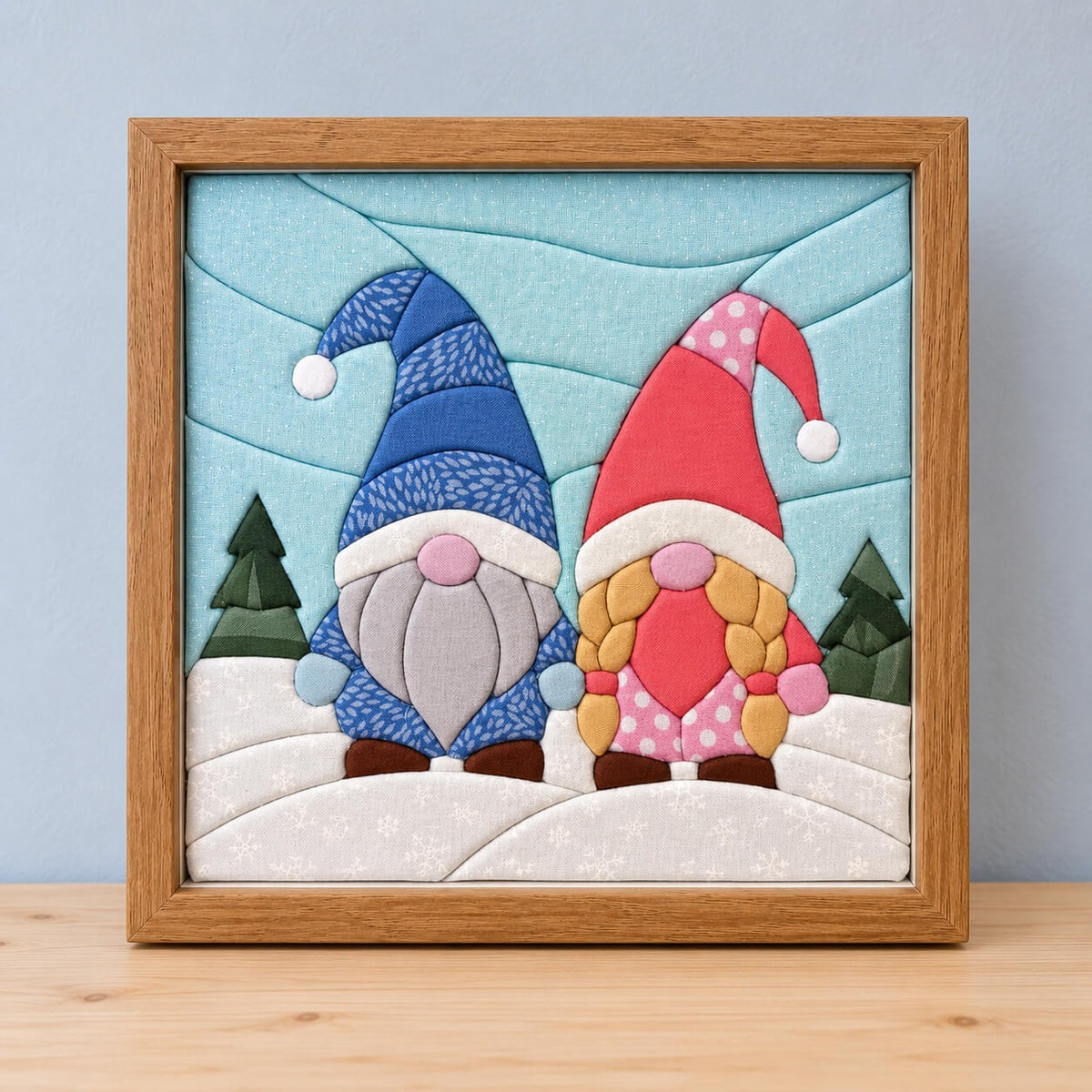 A square kinusaiga piece in a natural wood frame, standing on a wooden shelf against a pale blue wall: two gnomes side by side on snowy hills, one in a tall blue patterned hat with a grey beard, the other in a pink hat with a polka-dot tip and blonde braids, between two small pine trees under a pale aqua sky.
