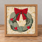 A Christmas wreath kinusaiga piece in a natural wood frame, standing on a wooden table against a grey wall: a ring of leaves in sage and deep green floral prints, a red bow at the top, and three clusters of round red holly berries, on a cream background printed with gold swirls and stars.