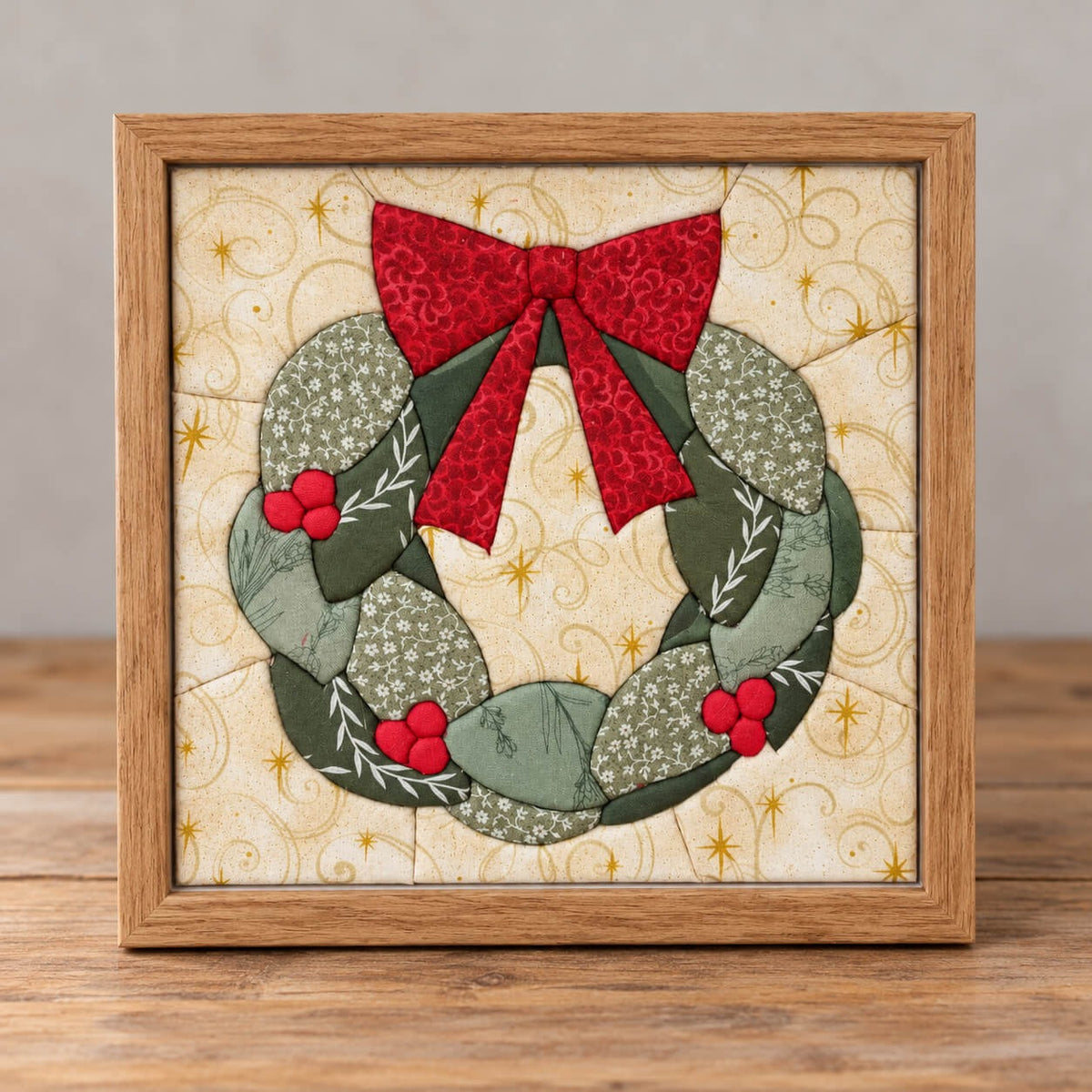 A Christmas wreath kinusaiga piece in a natural wood frame, standing on a wooden table against a grey wall: a ring of leaves in sage and deep green floral prints, a red bow at the top, and three clusters of round red holly berries, on a cream background printed with gold swirls and stars.