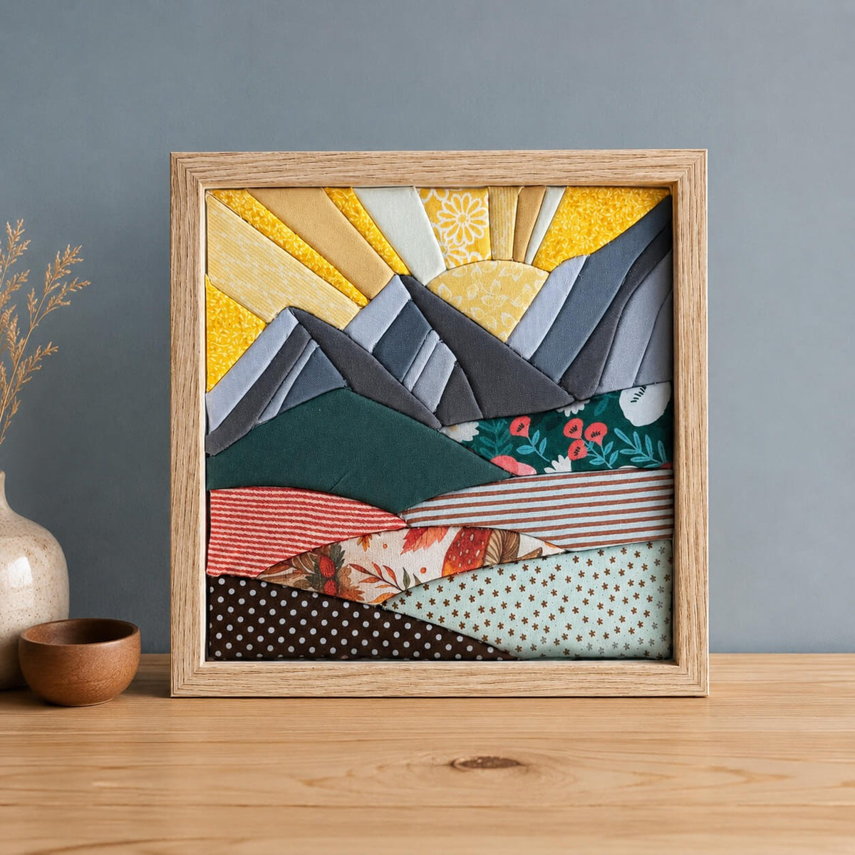 A square kinusaiga wall piece in a natural wood frame, standing on a wood table beside a speckled vase and a small wooden bowl: a grey and slate mountain range with a yellow sun and rays behind it, over layered hills of green, red stripe, floral and polka dot fabric.