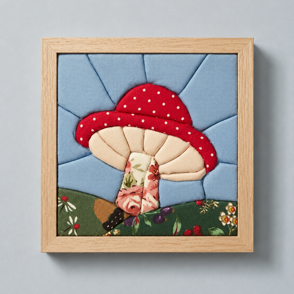 A framed square kinusaiga wall hanging featuring a whimsical mushroom design. The craft shows a red mushroom with white polka dots on a light blue background, with a floral stem and a green grassy base.