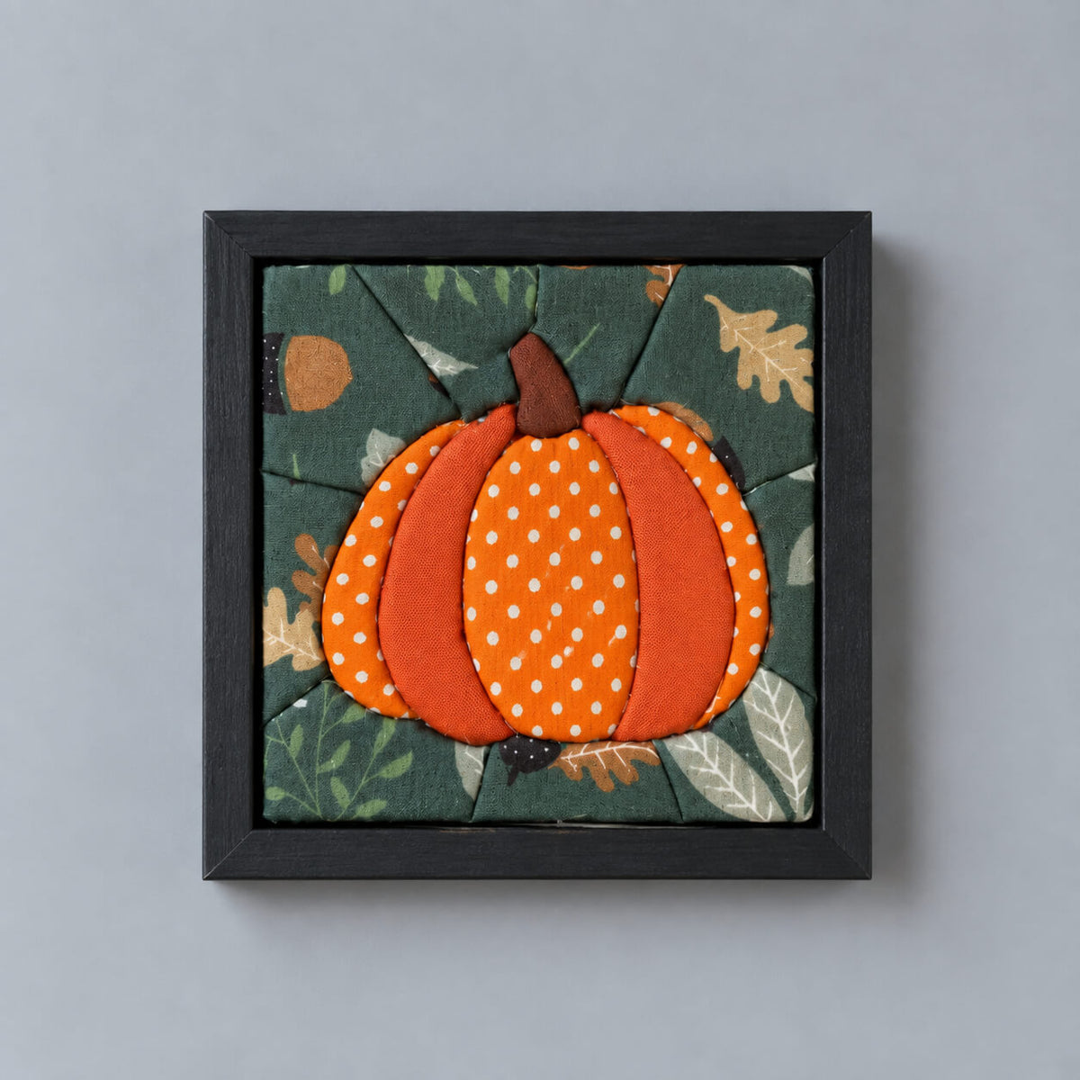 A square framed fabric wall art piece featuring a stylized orange pumpkin with white polka dots on a dark green autumn leaf patterned background, mounted on a light gray wall.