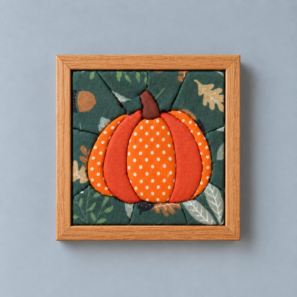 A square framed fabric wall hanging featuring a stylized orange pumpkin with white polka dots on a dark green autumn leaf background, set in a light wood frame against a solid gray wall.