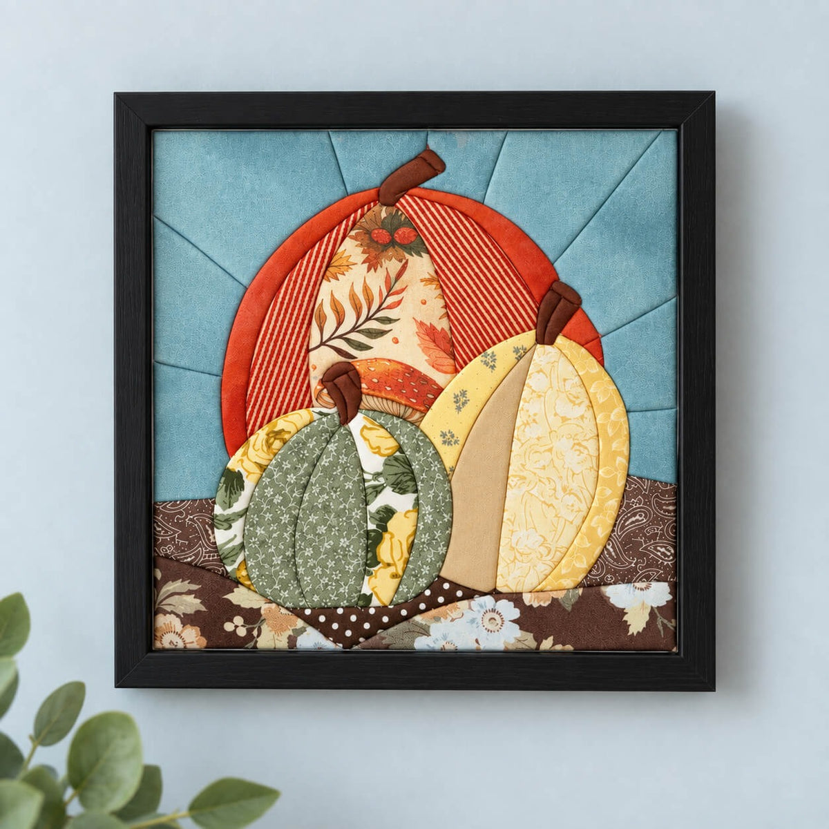 The same three-pumpkin design in a black frame, hanging flat on a pale grey wall.