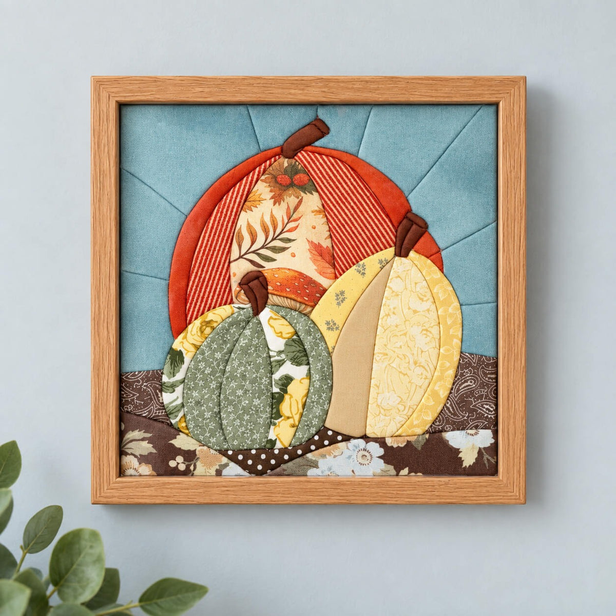 A square kinusaiga wall piece in a natural wood frame, hanging flat on a pale grey wall: three pumpkins in orange stripe, sage green floral and yellow fabric, stems tucked in brown, against a blue sky with rays, over rolling ground of brown paisley, polka dot and floral prints.