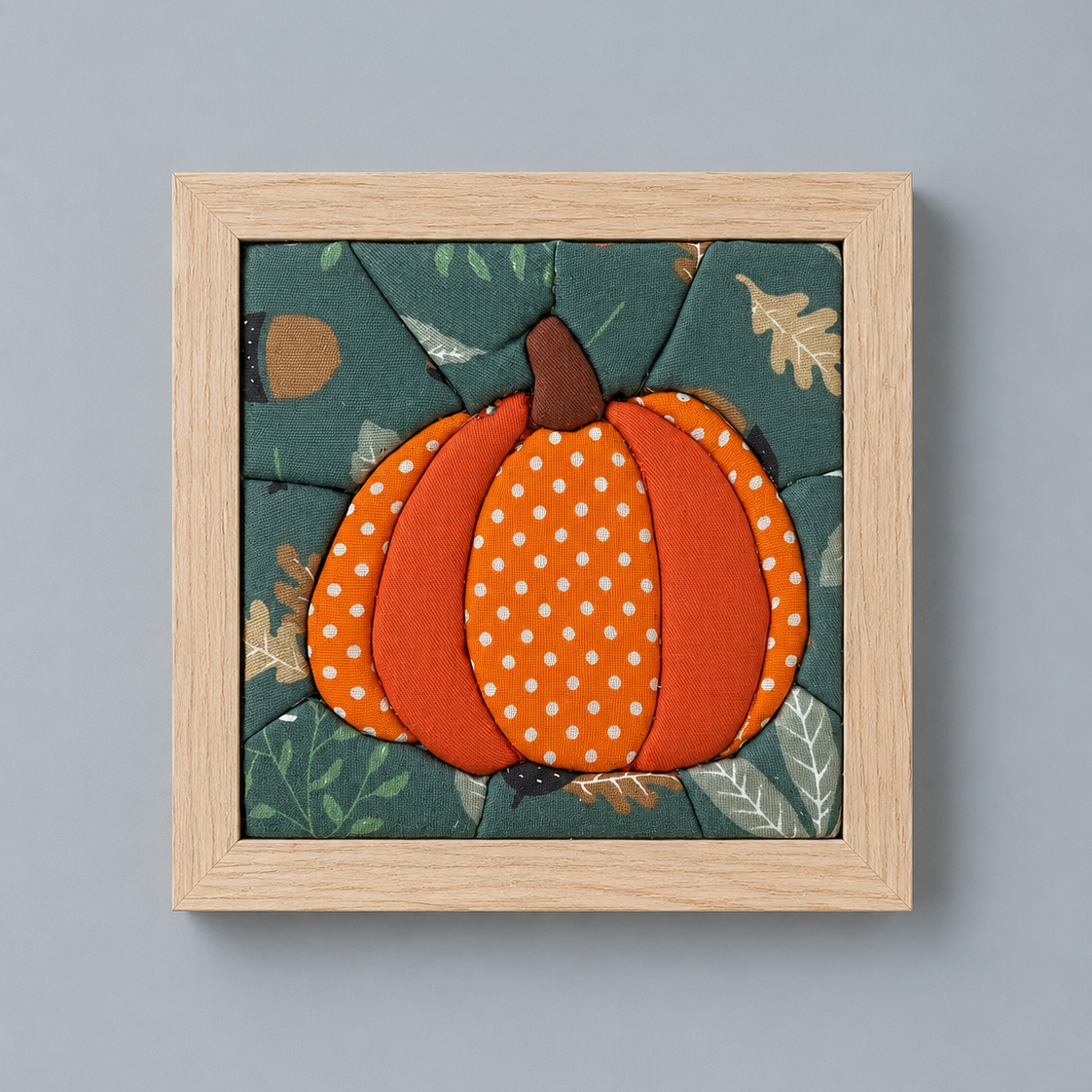 A square framed wall art piece featuring a stylized orange pumpkin with white polka dots on a dark teal autumn background with leaves, set in a light natural wood frame against a solid gray wall.