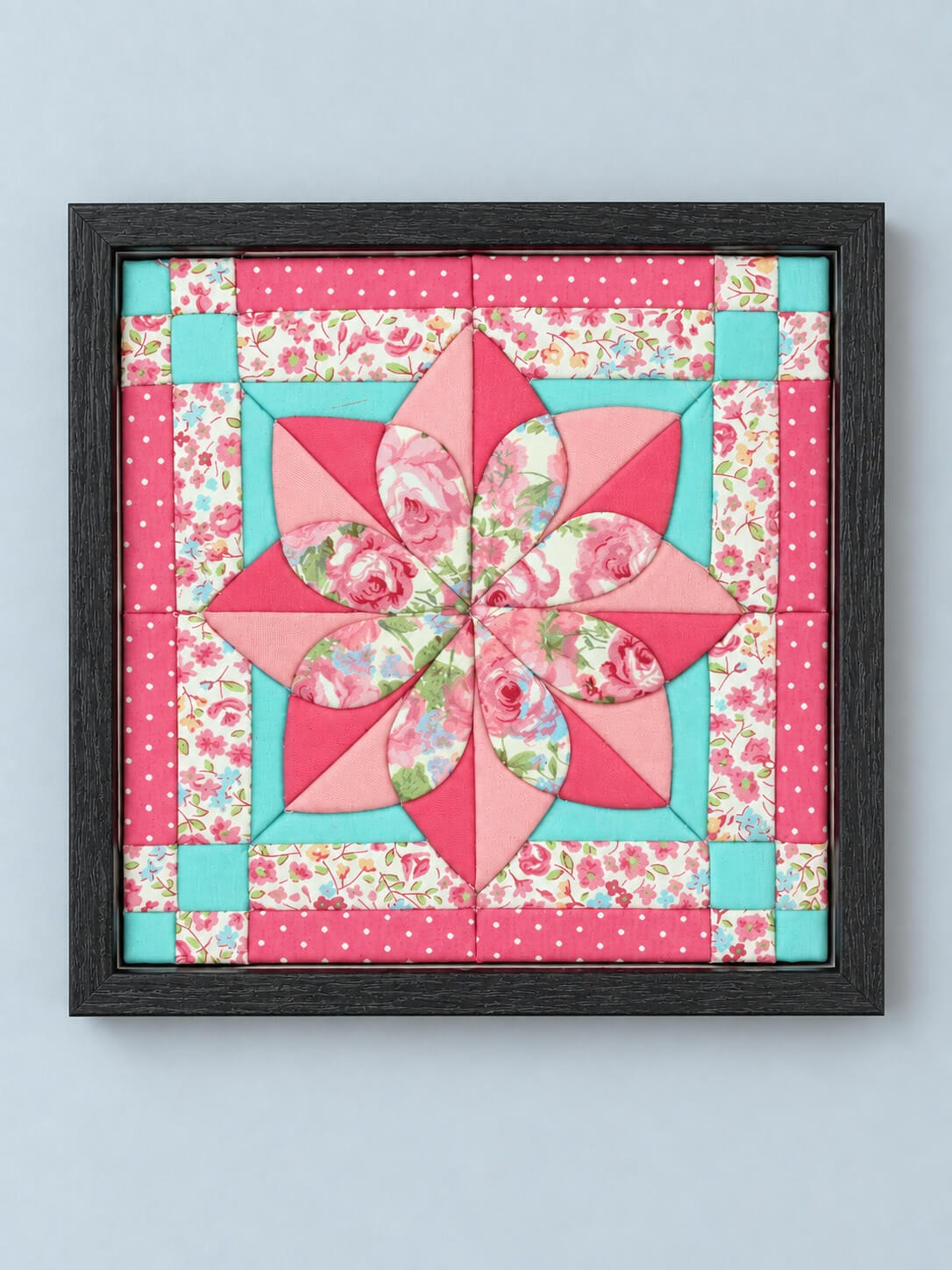 The same quilt flower design in a black frame, hanging flat against a pale grey wall.