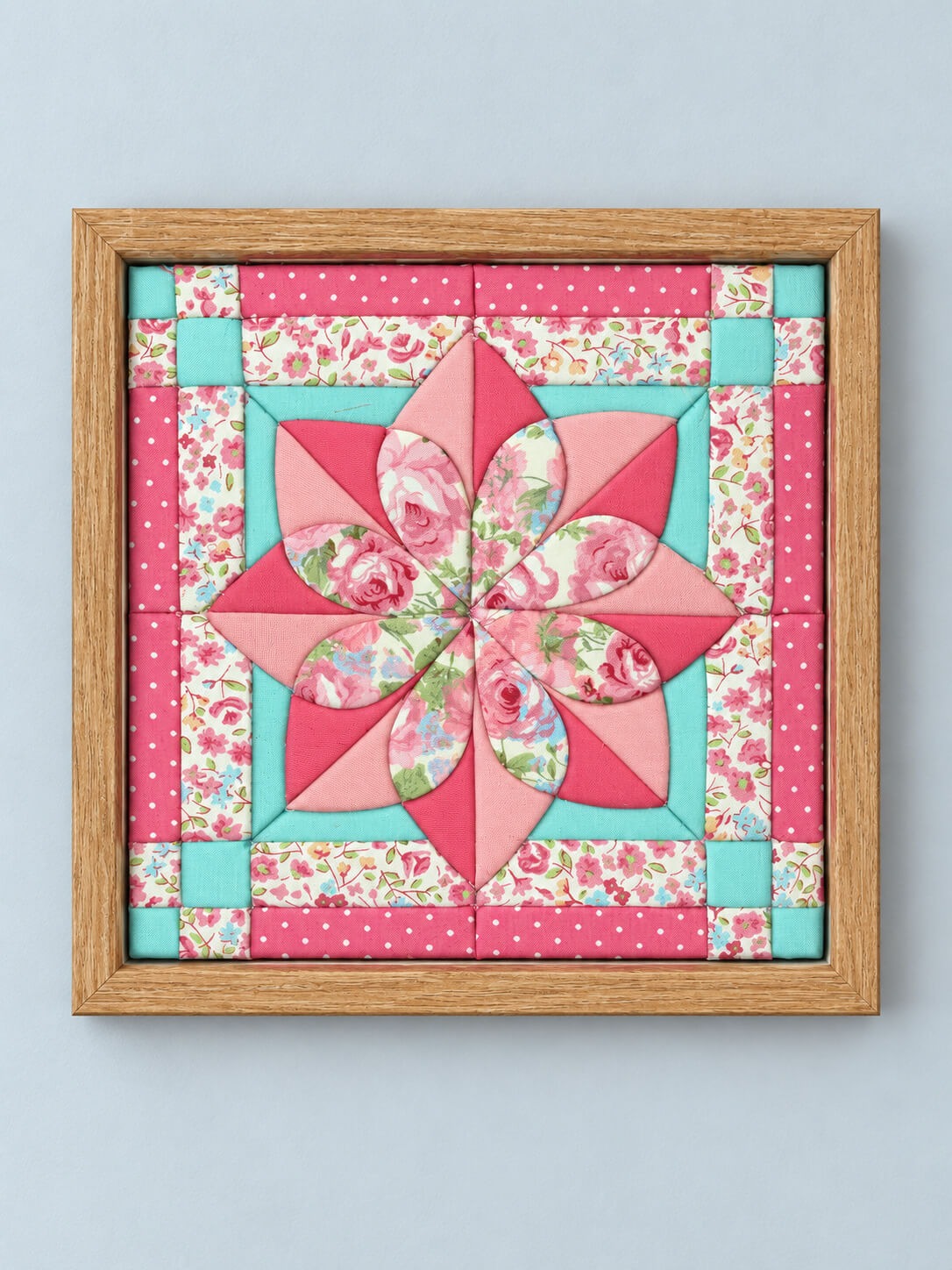 A square kinusaiga wall piece in a natural wood frame: an eight-petal quilt-block flower in pinks and rose florals on aqua, with a polka dot and floral border.