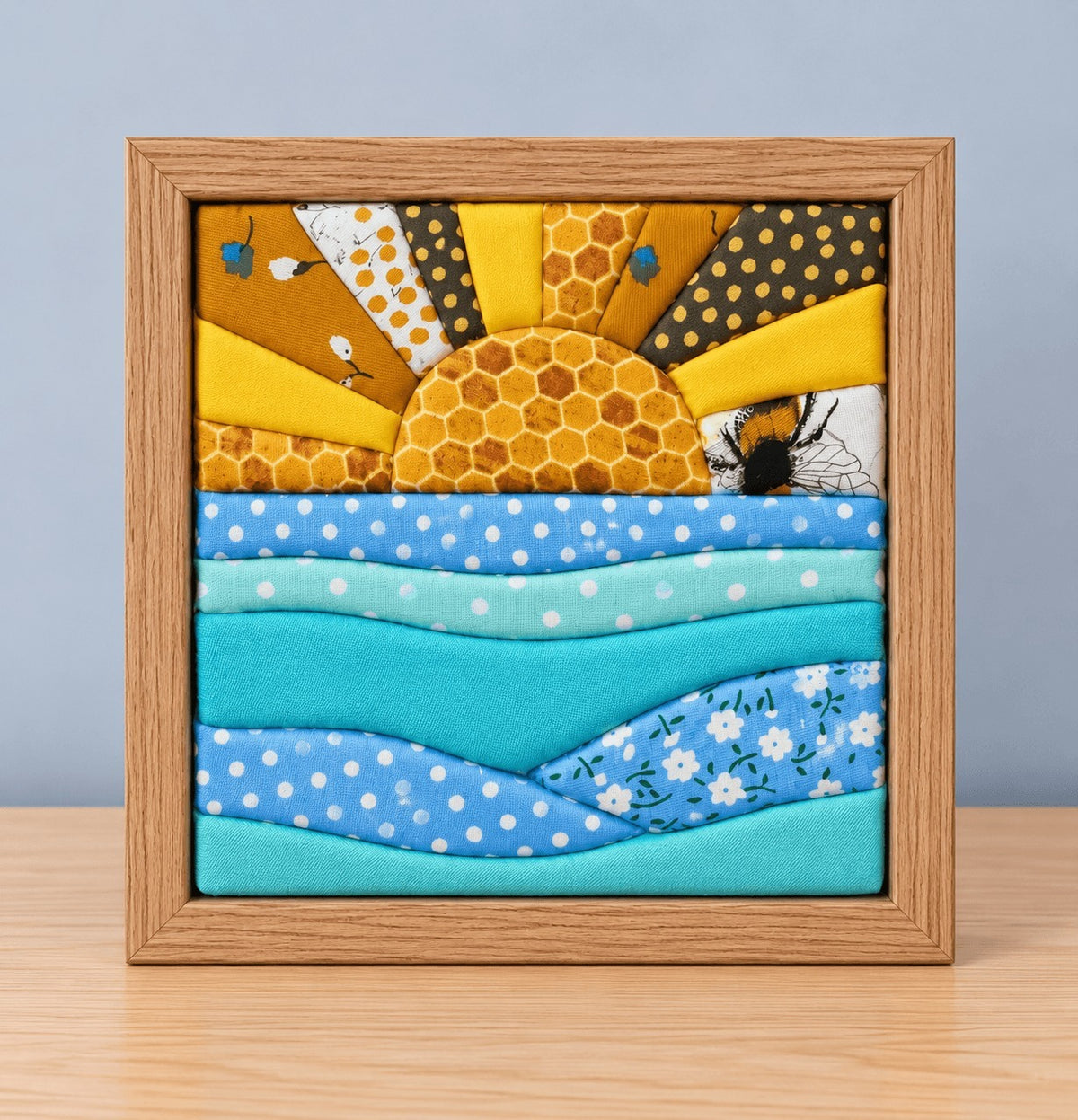 A square kinusaiga wall piece in a natural wood frame, standing on a light wood table against a grey-blue wall: a honeycomb sun rising with rays in yellows, mustard florals and gold polka dots, a bee in the corner, over waves of blue and turquoise polka dot, solid and floral fabric.
